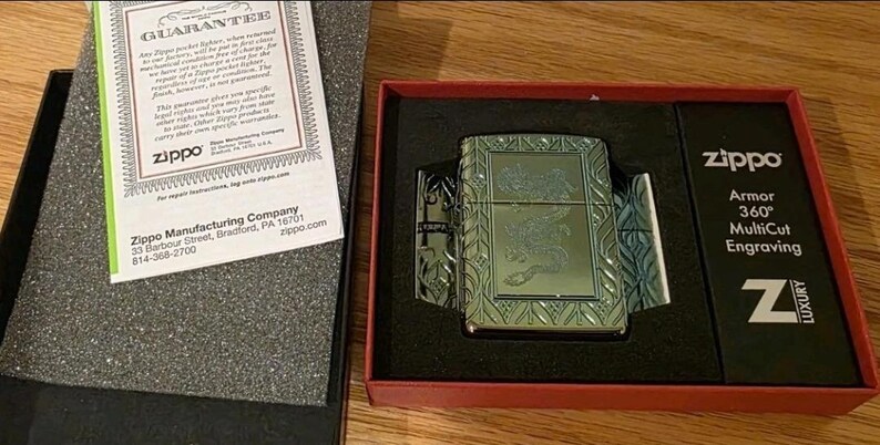 K&ouml;nnte beinhalten: Ein silbernes Zippo-Feuerzeug mit eingraviertem Design, pr&auml;sentiert in einer rot-schwarzen Box. Die Box enth&auml;lt eine Garantie und den Text "Armor 360&deg; MultiCut Engraving". Das Feuerzeug ist ein Luxusartikel.