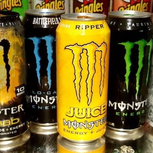 Puede incluir: Una colección de latas de bebidas energéticas Monster en varios colores, incluyendo naranja, amarillo, negro y plateado. Cada lata presenta el logotipo de la garra de Monster y nombres de productos como "Rehab", "Juice" y "Zero Ultra". Las latas están sobre una superficie reflectante.
