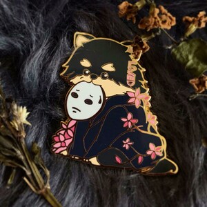 Puede incluir: Pin de esmalte dorado con un perro de dibujos animados en blanco y negro que lleva un kimono azul con flores de cerezo rosas y sostiene una m&aacute;scara blanca con una cara triste. El perro lleva un collar de oro con la palabra "Loved" escrita en &eacute;l.