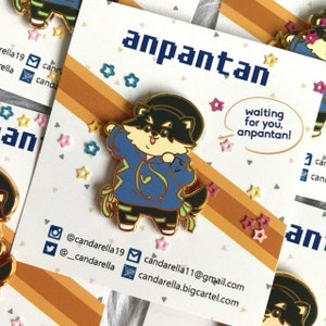 Puede incluir: Un pin de esmalte dorado con un perro de dibujos animados que lleva una sudadera azul con un dise&ntilde;o de rayo verde. El perro lleva un sombrero negro y tiene una nariz y una boca negras. El pin est&aacute; sobre un fondo marr&oacute;n y naranja con el texto "anpantan" y "waiting for you, anpantan!" en blanco. El pin est&aacute; rodeado de peque&ntilde;as estrellas rosas, amarillas y azules.