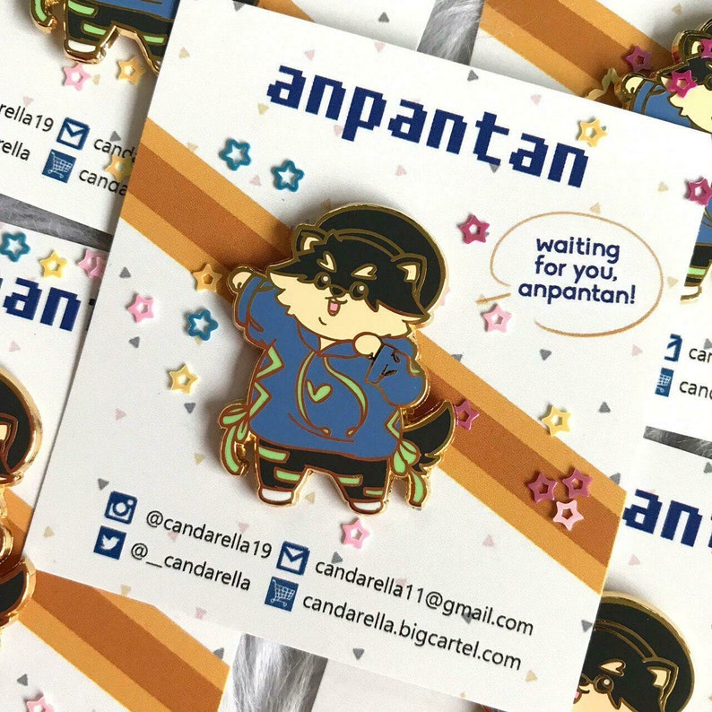 Puede incluir: Un pin de esmalte dorado con un perro de dibujos animados que lleva una sudadera azul con un dise&ntilde;o de rayo verde. El perro lleva un sombrero negro y tiene una nariz y una boca negras. El pin est&aacute; sobre un fondo marr&oacute;n y naranja con el texto "anpantan" y "waiting for you, anpantan!" en blanco. El pin est&aacute; rodeado de peque&ntilde;as estrellas rosas, amarillas y azules.