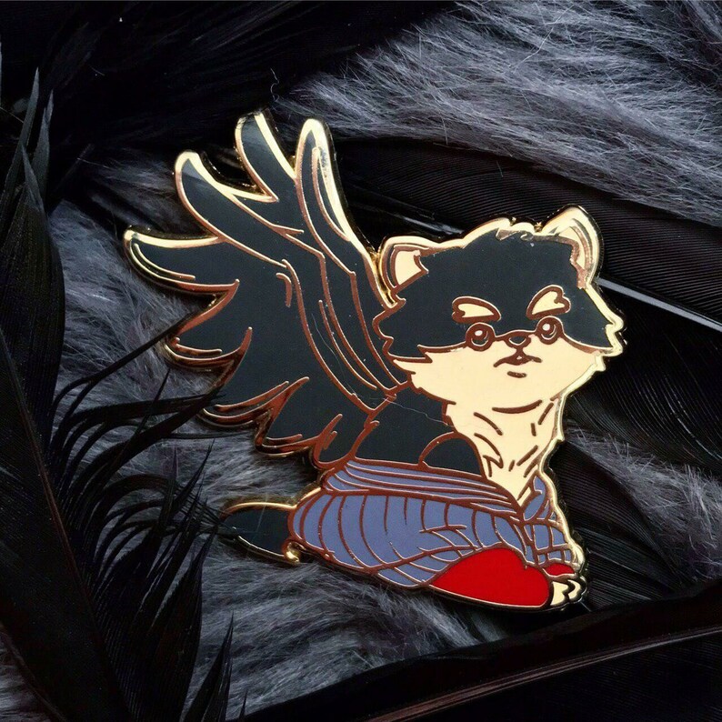 Puede incluir: Un pin de esmalte dorado con un perro blanco y negro con alas. El perro lleva un atuendo morado y rojo. El pin est&aacute; colocado sobre un fondo negro y gris.