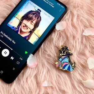 Puede incluir: Un tel&eacute;fono inteligente negro que muestra una aplicaci&oacute;n de reproductor de m&uacute;sica con la canci&oacute;n "It's Definitely You" de Jin en reproducci&oacute;n. La portada del &aacute;lbum presenta a un hombre con un atuendo tradicional coreano. Un pin de esmalte dorado de un personaje de dibujos animados con un atuendo azul y rosa con una espada se encuentra sobre una superficie rosada y peluda.