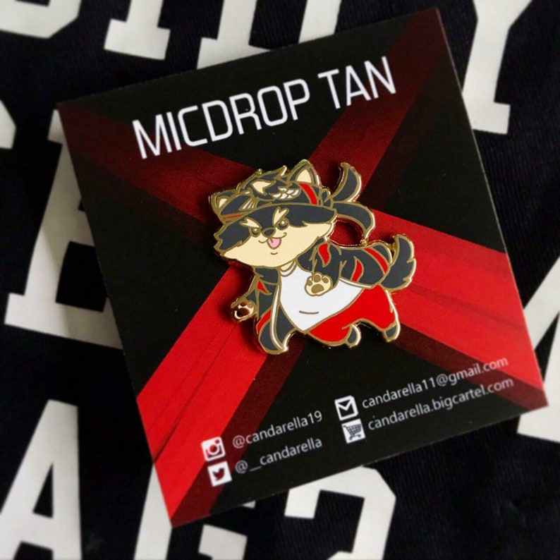 Puede incluir: Un pin de esmalte dorado con un mapache de dibujos animados que lleva una chaqueta negra y roja, una camisa blanca y pantalones rojos. El mapache lleva una diadema negra con una estrella dorada. El pin est&aacute; sobre un fondo rojo y negro con el texto "MICDROP TAN" en letras doradas. El texto "@candarella19", "@candarella", "candarella11@gmail.com" y "candarella.bigcartel.com" tambi&eacute;n es visible en el fondo.