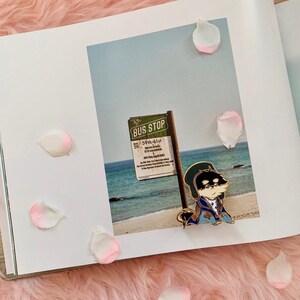 Puede incluir: Un pin de esmalte dorado con un personaje de dibujos animados que lleva una chaqueta azul y un sombrero negro. El personaje est&aacute; de pie frente a un cartel que dice "Bus Stop 59th-61st".