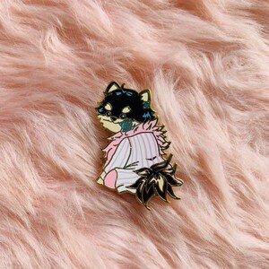 Puede incluir: Un pin de esmalte negro y blanco de un perro con un pijama de rayas rosa y azul con un cuello blanco esponjoso. El perro est&aacute; sentado con la cola enroscada detr&aacute;s de &eacute;l.