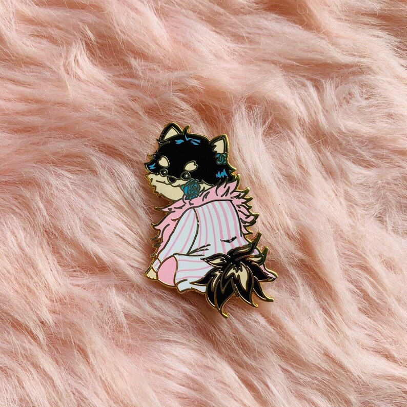 Puede incluir: Un pin de esmalte negro y blanco de un perro con un pijama de rayas rosa y azul con un cuello blanco esponjoso. El perro est&aacute; sentado con la cola enroscada detr&aacute;s de &eacute;l.