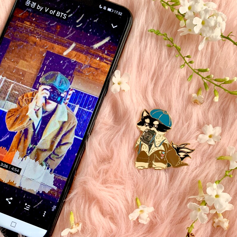 Puede incluir: Un pin de esmalte dorado con un perro de dibujos animados que lleva una gorra azul y sostiene una c&aacute;mara. El perro lleva una chaqueta marr&oacute;n y tiene un hocico blanco. El pin est&aacute; sobre un fondo rosa peludo.