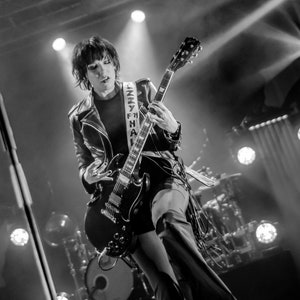 May include: Black and white image of a person playing a black electric guitar on stage. The guitar strap reads "Jenny Hall". The person is wearing a black leather jacket and platform boots. Stage lights illuminate the scene.