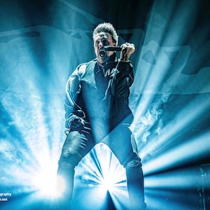 Peut inclure: Photographie d'un concert en direct, avec un chanteur sur scène, éclairé par des projecteurs bleus. Le chanteur tient un microphone et chante la bouche ouverte. L'arrière-plan est sombre, avec un logo et une adresse web visibles.