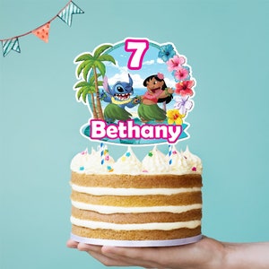 Puede incluir: Un pastel en capas con glaseado blanco y dos velas. El pastel est&aacute; cubierto con un adorno colorido con los personajes Stitch y Lilo, el n&uacute;mero 7 y el nombre "Bethany". El fondo es azul claro.