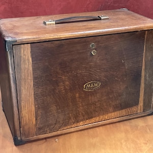 May include: A vintage wooden box with a dark brown finish, leather handle and metal corner reinforcements. The box has a front door with a keyhole and a small brass emblem. The logo "M&W" is visible on the front.