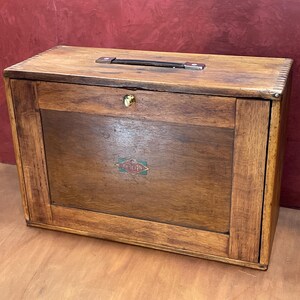 May include: A wooden box with a handle and a lock. The box is made of dark brown wood and has a rectangular shape. A small, colourful emblem is on the front of the box. The box is sitting on a wooden surface.