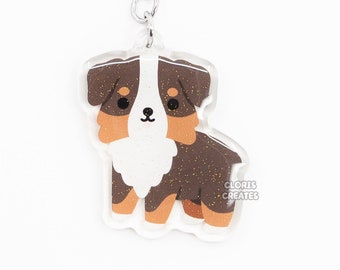 May include: A brown, white, and black Australian Shepherd dog keychain with glitter accents. The keychain is made of clear acrylic and has a silver metal key ring.