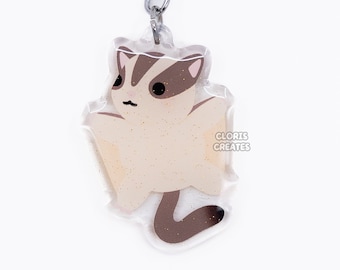 May include: A beige and brown sugar glider keychain with a silver key ring. The keychain is made of clear acrylic and has a glittery finish. The sugar glider is depicted in a flying pose with its wings outstretched. The text "GLORIES CREATES" is printed on the keychain.