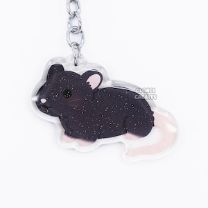 Puede incluir: Un llavero de rata de purpurina negra y blanca con un llavero plateado. La rata está en posición sentada con la cola enroscada detrás de ella. El llavero está hecho de acrílico transparente.