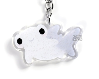 May include: A white glitter hammerhead shark keychain with black eyes. The keychain is attached to a silver metal key ring.