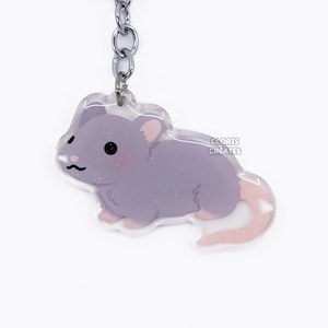 Puede incluir: Un llavero de rata morado con una cola rosa y un llavero plateado. La rata tiene una cara de dibujos animados linda con ojos negros y una nariz rosa.