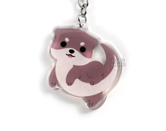May include: A brown and white otter keychain with a silver key ring. The otter is depicted in a cartoon style with a cute expression. The keychain is made of clear acrylic and has a glossy finish.