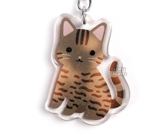 May include: A brown and black tabby cat keychain with a silver key ring. The cat is sitting with its tail curled around its legs. The keychain is made of clear acrylic.