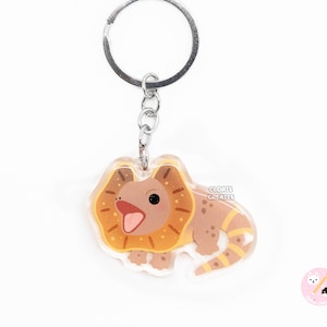 Puede incluir: Un llavero de lagarto con volantes marrón y amarillo con un llavero plateado. El lagarto es de estilo de dibujos animados y tiene un gran volante alrededor del cuello. El texto "CLORIS CREATES" está impreso en el llavero.