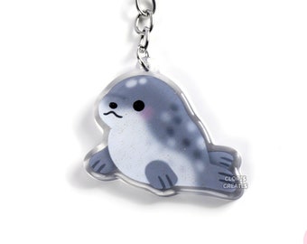 May include: A gray and white seal keychain with a silver chain and ring. The seal is depicted in a cartoon style with a cute expression. The keychain is made of clear acrylic and has a glossy finish.
