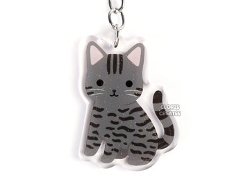 May include: A grey and black tabby cat keyring made of clear acrylic with a silver metal key ring. The keyring features a cute cartoon cat with black stripes and a white belly. The text "GLORIE CREATES" is printed on the bottom right corner of the keyring.