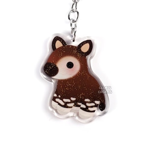 May include: A brown and white okapi keychain with glitter accents. The keychain is made of clear acrylic and has a silver metal ring.