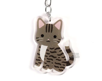 May include: A brown and white tabby cat keychain with a silver key ring. The cat is sitting with its tail curled around its legs. The keychain is made of clear acrylic.