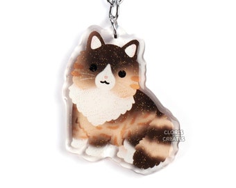 Pode incluir: Um chaveiro de acrílico transparente com um gato de desenho animado com pelo castanho, branco e bege. O gato está sentado. O chaveiro tem um anel e uma corrente de metal prateado. O texto "CLORIS CREATES" é visível.
