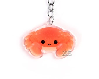 May include: A red and orange cartoon crab keyring with a smiling face. The keyring is made of clear acrylic and has a silver metal chain.