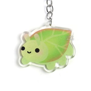 Puede incluir: Un llavero de dibujos animados de un bicho de hoja verde y rosa con una cadena y un anillo plateados. El llavero tiene el texto "Cloris Creates".