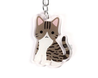 May include: A brown and white tabby cat keyring with a silver key ring. The cat is sitting with its tail curled around its legs. The keyring is made of clear acrylic.