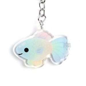 May include: A clear acrylic keychain featuring a pastel rainbow-colored fish design with a silver keyring and chain. The fish has a smiling face and the text "GLORIE CREATES" is visible on the right side of the fish.