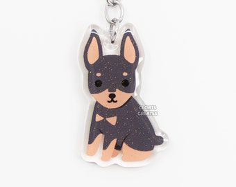 May include: A black and tan miniature pinscher dog keychain with glitter accents. The keychain is made of clear acrylic and has a silver metal ring.