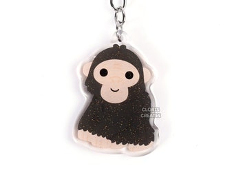 May include: A black and brown glitter keychain featuring a cartoon ape with a smiling face. The keychain is attached to a silver metal ring and chain.