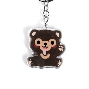 May include: A brown bear keyring with a glittery finish. The bear is sitting with its tongue sticking out and one paw raised. The keyring is attached to a silver chain and ring.