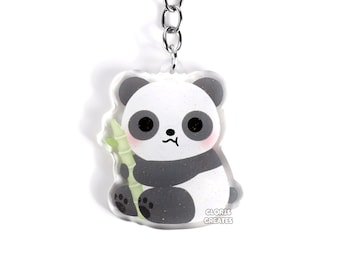 Peut inclure: Un porte-clés en acrylique transparent représentant un panda dessin animé tenant un morceau de bambou. Le panda est noir et blanc avec une expression mignonne. Le porte-clés est attaché à une chaîne argentée et un anneau métallique noir. Le texte "CLORIS CREATES" est imprimé sur le porte-clés.