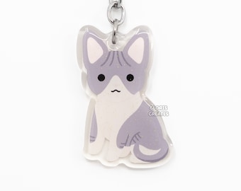 Pode incluir: Um chaveiro de gato roxo e branco com um chaveiro prateado. O gato está sentado com o rabo enrolado em torno de suas pernas. O chaveiro é feito de acrílico transparente e tem um acabamento brilhante. O texto "GLORIS CREATES" é impresso na parte inferior do chaveiro.