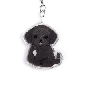 May include: A black and glittery dog-shaped keychain with a silver keyring and chain. The dog has a white spot and a pink tongue. The keychain has the text "CLORIS CREATES" on the side.