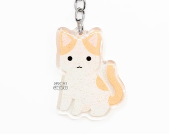 May include: A clear acrylic keychain featuring a glittery white and orange cat design. The cat has black eyes and a small black mouth. The keychain has a silver metal chain and ring. The text "GLORIS CREATES" is printed on the keychain.