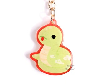 May include: A green snake with a red tongue is curled up on a red background. The snake is a keychain with a gold chain and ring. The keychain is labelled "Gloomies Creates".