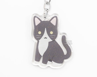 May include: A gray and white cat keychain with glitter accents. The keychain is made of clear acrylic and features a silver metal chain and ring.