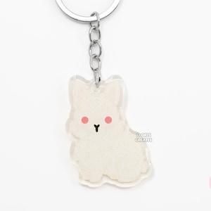 May include: White glitter dog keychain with pink cheeks and a black nose. The keychain is attached to a silver metal ring and chain. The text "CLORIS CREATES" is printed on the bottom right corner of the keychain.