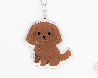 May include: A brown glitter keychain featuring a cartoon illustration of a poodle dog. The keychain is attached to a silver metal ring and chain.