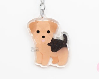 Puede incluir: Un llavero de acrílico transparente con un Yorkshire Terrier de dibujos animados con pelaje marrón, orejas negras y cola negra. El perro está de pie sobre sus cuatro patas y tiene la lengua rosa que sobresale. El llavero está unido a un anillo y una cadena de metal plateado.