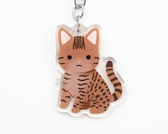 May include: A brown and black tabby cat keychain with a silver key ring. The cat is sitting with its tail curled around its legs. The keychain is made of clear acrylic.