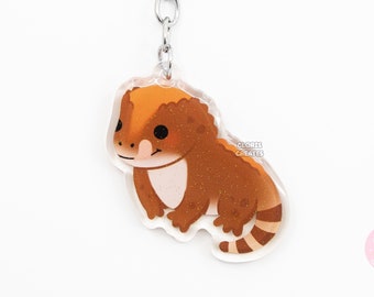 May include: A brown and orange bearded dragon keychain with a silver key ring. The keychain is made of clear acrylic and features a cartoon illustration of a bearded dragon.