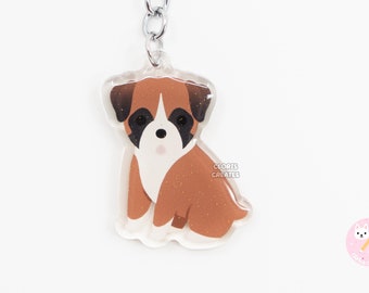 Puede incluir: Un llavero de acrílico transparente con una ilustración de dibujos animados de un cachorro boxer marrón y blanco. El llavero tiene un anillo y una cadena de metal plateado.