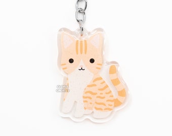 May include: Orange and white tabby cat keychain with a silver key ring. The cat is sitting with its tail curled around its legs. The keychain is made of clear acrylic and has a glitter finish.